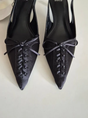 Svarta spetsiga pumps från MNG - Säljer ett par eleganta svarta pumps från MNG med spetsig tå, låg klack och öppen häl. Skorna har dekorativ snörning och rosett framtill samt är tillverkade i ett glansigt satinliknande material. Perfekta för dig som gillar en chic och trendig look. Med box.