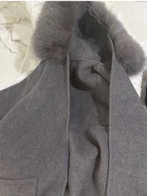 Grå kappa med rävpäls - Stilren grå kappa med stor huva och mjuk rävpäls runt kragen. Kappan har raka ärmar och en klassisk passform, perfekt för kyliga dagar. Materialet ser ut att vara ullblandning för extra värme och komfort. Köpt för ca 4000kr (xs men passar även s)