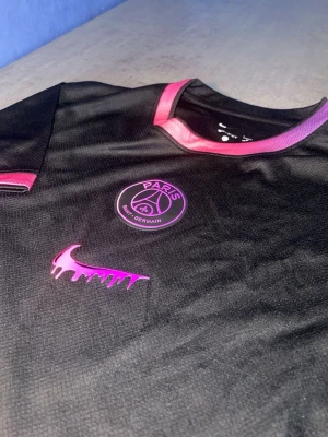 Svart PSG x Nike t-shirt med rosa detaljer - Säljer en svart PSG t-shirt från Nike med rosa detaljer på krage och ärmslut. Tröjan har PSG-logga och en unik rosa Nike swoosh på bröstet. Tillverkad i ett lätt och ventilerande material, perfekt för träning eller streetwear.