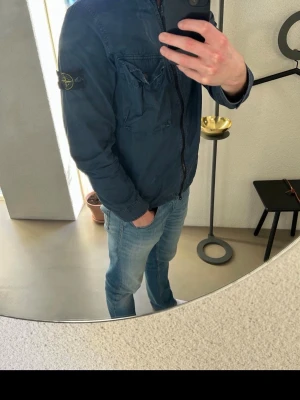 Stone Island overshirt - Stone Island jacka för herr i storlek S. Jackan är blå och tillverkad i bomull. Modellen har dragkedja framtill, bröstficka med lock samt sidofickor. Ärmarna har knapp vid mudd och den klassiska Stone Island-detaljen syns på ärmen. Passar som lätt ytterjacka i vardagen. Nypris ca. 4500kr.