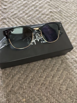 Ray-Ban  solglasögon - Snygga Ray-Ban Clubmaster solglasögon med svart ovandel och guldfärgade detaljer. Klassisk retroform med mörka glas och metallbåge. Kommer med originalfodral, putsduk och box. Perfekta för dig som vill ha en tidlös och trendig look.