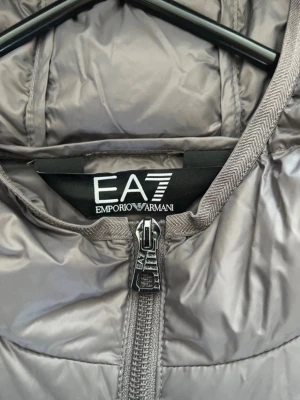 Grå pufferjacka från EA7 Emporio Armani - Snygg grå pufferjacka från EA7 Emporio Armani med huva och dragkedja framtill. Jackan har ett quiltat mönster och EA7-logga på bröstet. Perfekt för kalla dagar och ger en clean, modern look. Tillverkad i lätt och värmande material.