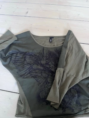 y2k topp - Säljer en olivgrön topp med stort svart tryck framtill. Toppen har bred halsringning, trekvartsärmar och en loose passform. Toppen är köpt på sellpy💓storlek s men passar de flästa ioch med att den sitter löst