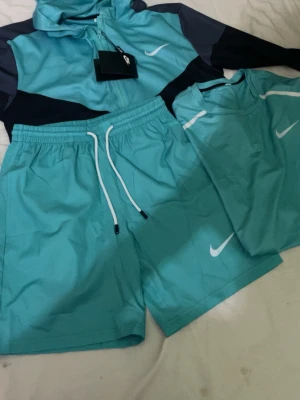 Nike Miller set  - Snyggt träningsset från Nike i turkos med vita detaljer. Setet består av en långärmad zip-tröja, t-shirt och shorts. Alla plagg har Nike-loggan tryckt på bröstet eller benet. Materialet är lätt och snabbtorkande, perfekt för träning eller sport.