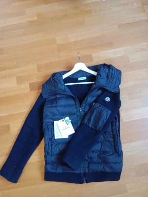Svart pufferjacka från Moncler - Snygg svart pufferjacka från Moncler med stickade ärmar och huva. Jackan har dragkedja framtill, fickor med dragkedja och en Moncler-logga på ärmen. Materialmix med quiltad front och stickade detaljer gör den både trendig och varm. Passar 170-180 men kan gå för större. Pris inte hugget i sten! Fraktar inom 12 timmar.
