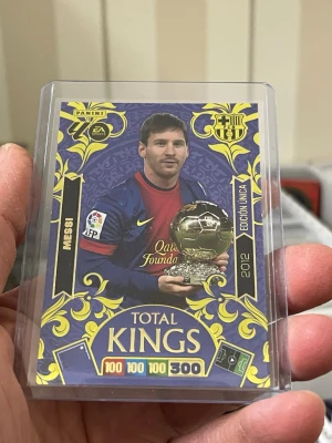 Panini Adrenalyn XL Total Kings kort Lionel Messi - Samlarbild från Panini Adrenalyn XL-serien, Total Kings, med färgstark design i blått och guld. Kortet visar en fotbollsspelare med Ballon d'Or-trofé och har detaljer som klubbmärke, årtal 2012 och spelarnamn. Baksidan har Adrenalyn XL-logga och kod.