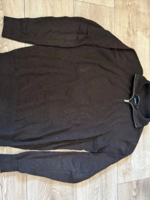 Brun stickad tröja med halv dragkedja - Mörkbrun stickad tröja från S.C.W med halv dragkedja och hög krage. Tillverkad i återvunnet material. Tröjan har långa ärmar och ribbade muddar vid ärmslut och nederkant. Perfekt för lager på lager och en clean look.