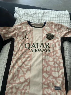 PSG x Jordan matchtröja beige/rosa - Säljer en PSG x Jordan matchtröja i beige och ljusrosa med svart logga och detaljer. Tröjan har ett unikt mönster, svart Paris Saint-Germain-märke på bröstet och Qatar Airways-tryck framtill. Tillverkad i lätt och ventilerande material, perfekt för fotboll eller träning.