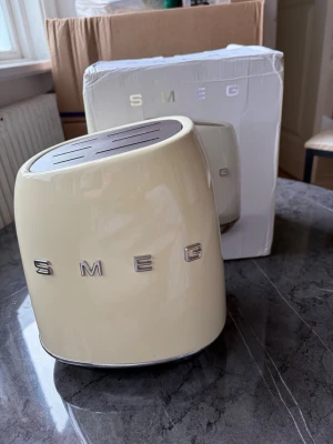 Krämfärgad knivblock från Smeg - Snyggt knivblock från Smeg i krämfärgad nyans med retroinspirerad design. Tillverkat i robust material med plats för flera knivar. Passar perfekt på köksbänken och ger ett stilrent intryck. Smeg-logga i silver på framsidan. Kvitto finns 