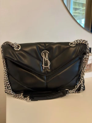 Svart quiltad handväska från Steve Madden - Snygg svart quiltad handväska från Steve Madden med silverfärgad kedjerem och logga framtill. Väskan har en mjuk, vadderad look och V-formade sömmar. Perfekt för dig som gillar trendiga accessoarer med lite edge.