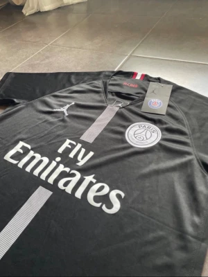 Svart PSG matchtröja Jordan x Fly Emirates - Snygg svart PSG matchtröja i samarbete med Jordan och Fly Emirates. Tröjan har korta ärmar, rund hals och en grå vertikal rand på framsidan. PSG-logga och Jumpman-logga på bröstet. Tillverkad i lätt och ventilerande material, perfekt för fotboll eller träning.