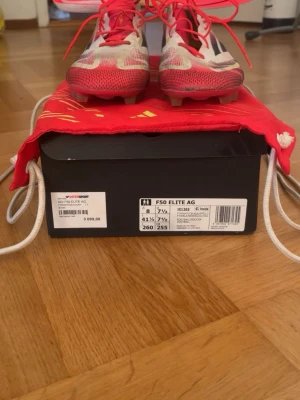 Adidas F50 Elite AG fotbollsskor - Säljer ett par Adidas F50 Elite AG fotbollsskor i vitt, rött och svart. Skorna har en modern, strömlinjeformad design med röda snören och detaljer. Ovandelen är i syntetmaterial för lätt vikt och bra bollkontroll. Perfekta för spel på konstgräs. Innersulor och röd påse medföljer.