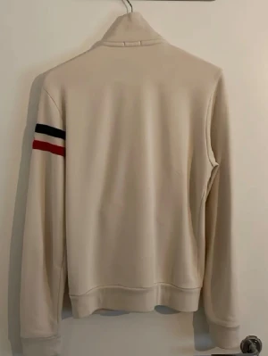  zip hoodie  Moncler - Snygg beige hoodie från Moncler med dragkedja framtill och två fickor. På vänster ärm finns Monclers logga och en rand i blått, vitt och rött. Hoodien har hög krage och ribbade muddar vid ärmslut och nederkant. Perfekt för en clean och stilren look. Självklart så är den äkta 