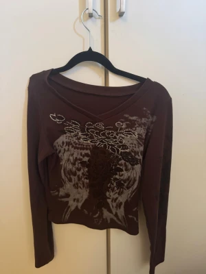 Brun långärmad topp med mönster - Brun långärmad topp med v-ringning och dekorativa detaljer framtill i form av blommigt mönster och glittriga stenar. Snygg passform och perfekt för dig som gillar unika prints och lite extra bling.