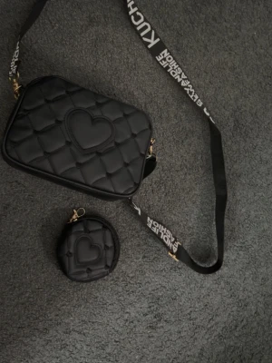 Svart axelväska med hjärtdetalj - Säljer en svart axelväska med quiltat mönster och en stor hjärtdetalj på framsidan. Väskan har en bred axelrem med vit text och matchande liten rund coin purse med hjärtdetalj. Perfekt för dig som gillar trendiga accessoarer.
