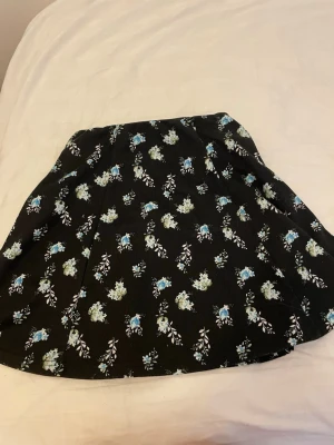 Svart blommig kortkjol - Snygg svart kortkjol med blommigt mönster i blått, vitt och grönt. Kjolen har en lätt utställd form och är perfekt för dig som gillar en feminin och trendig stil. Passar till både sneakers och boots för en cool look.