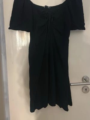 Svart offshoulder klänning med puffärm - Trendig svart offshoulder klänning med puffärmar och snygg snörningsdetalj framtill. Klänningen har en smickrande passform och är perfekt för dig som vill ha en enkel men stilren look. Tillverkad i mjukt tyg som känns skönt mot huden.