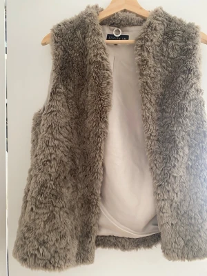 Beige fluffig pälsväst STOCKH LM - Supersnygg beige pälsväst från STOCKH LM i storlek XS/S. Västen är gjord i mjuk fuskpäls och har en öppen design utan knäppning. Perfekt att slänga över en hoodie eller tröja för en trendig lager-på-lager-look.