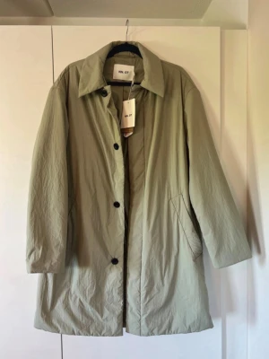 Ljusbeige trenchcoat från NN07 - Stilren ljusbeige trenchcoat från NN07 med klassisk krage, raka ärmar och knäppning med svarta knappar. Jackan har två snedställda fickor och är tillverkad i ett lätt, strukturerat material som ger en modern look. Perfekt för dig som gillar minimalistisk och clean stil.