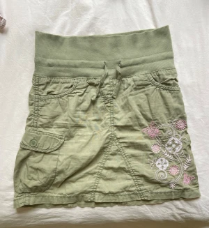 Y2k kjol - Snygg ljusgrön kortkjol med bred ribbad midja och snörning. Kjolen har flera fickor och ett broderat blommönster med rosa och vita detaljer framtill. Perfekt för vår och sommar, enkel att matcha till olika stilar.