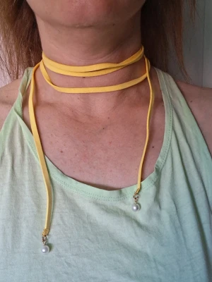 Gult chokerhalsband med pärlor - Snyggt gult chokerhalsband i mockaimitation som lindas flera varv runt halsen. Halsbandet har två hängande band med små vita pärlor längst ner, vilket ger en trendig och ungdomlig look. Perfekt accessoar för att lyfta din outfit.