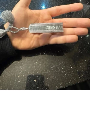 Grå håraccessoar från Dior - Säljer en snygg grå håraccessoar från Dior med texten 'Christian Dior J'Adior' broderad. Accessoaren är i flätad textil med dekorativa tofsar i ändarna, perfekt för att ge din frisyr en exklusiv touch.