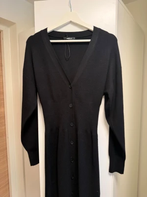 Svart lång stickad klänning Gina Tricot  - Svart lång stickad klänning från Gina Tricot med v-ringning och knappar hela vägen framtill. Klänningen har lång ärm och markerad midja för en snygg siluett. Perfekt till lager på lager och enkel att styla med accessoarer.