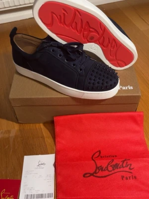Mörkblå Louboutin sneakers med nitar - Snygga svarta sneakers från Christian Louboutin med klassisk röd sula och svarta nitar på tån. Skorna har en låg profil, vita sulor och är tillverkade i mocka. Perfekta för dig som vill sticka ut med exklusiv design och edgy detaljer.