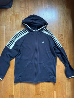 Svart Adidas hoodie med dragkedja - Klassisk svart hoodie från Adidas med dragkedja framtill och vita ränder längs ärmarna. Ikonisk Adidas-logga på bröstet och praktisk huva. Tillverkad i mjukt material, perfekt för en avslappnad streetwear-look.
