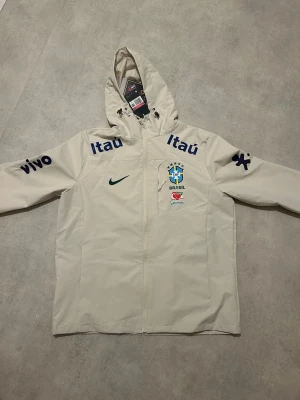 Vit Brasil Nike träningsjacka - Säljer en helt ny vit träningsjacka från Nike med Brasil landslagslogga på bröstet. Jackan har huva, dragkedja och flera sponsorloggor som Itaú och Vivo i blått. Perfekt för fotbollsträning eller sportiga dagar. Säljer den på grund av storlek. Vid snabb och smidigaffär kan jag sänka den lite billigare 