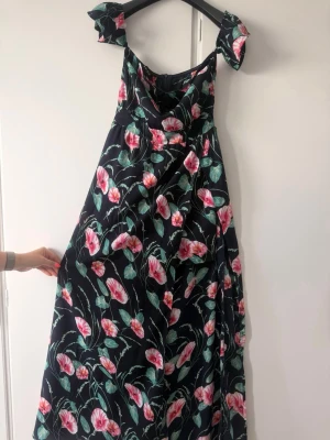 Blommig långklänning med rosettband - Svart långklänning med färgglatt blommönster i rosa, vitt och grönt. Klänningen har breda axelband med stora rosetter och en luftig, svepande passform. Perfekt för sommarens alla tillfällen.