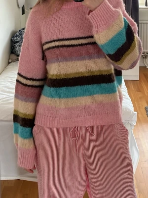 Rosa randig stickad tröja - Mysig stickad tröja i rosa med breda ränder i blått, brunt, gult, vitt och lila. Tröjan har rund halsringning, ribbade muddar och en avslappnad passform. Perfekt för dig som gillar färgglada och unika plagg.