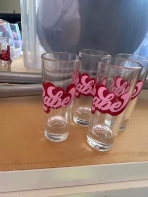 Set med shot glas med rosa tryck - Säljer ett set med fyra shot glas i transparent glas med rosa och röd text 'Babe' tryckt på framsidan. Glasen har en cylindrisk form och är perfekta för drycker. Trycket har en lekfull och ungdomlig design med hjärtformad bakgrund. 6cl