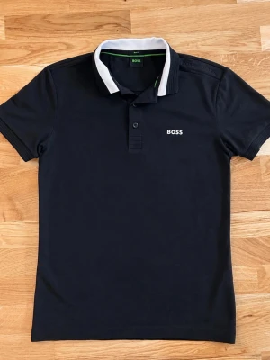 Svart pikétröja från BOSS - Klassisk svart pikétröja från BOSS med vit kontrastkrage och knappslå framtill. Tröjan har korta ärmar och BOSS-logga broderad på bröstet. Perfekt till både jeans och chinos för en clean look.