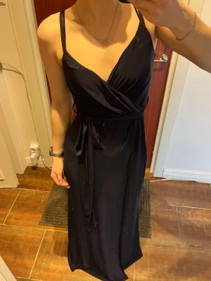 Marinblå omlottklänning med slits - Elegant marinblå långklänning med omlott över bysten, smala axelband och knytband i midjan. Klänningen har en hög slits på ena sidan som ger en snygg och modern look. Aldrig använd. 