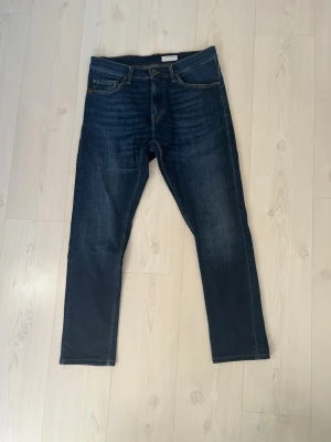 Mörkblå slim fit jeans - Snygga mörkblå slim fit jeans från  Tiger of Sweden storlek 33/34 med klassisk femficksdesign och kontrastsömmar. Jeansen har en rak passform och subtila slitningar framtill för en modern look. Perfekta till vardags och lätta att matcha med olika stilar. Uppsydda så lite kortare än 34