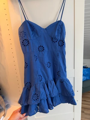 Blå kort klänning med volang - Söt blå kort klänning med tunna axelband och volangkant. Klänningen har broderade blommönster och är tillverkad i bomull. Perfekt för varma dagar och har en luftig, somrig känsla.