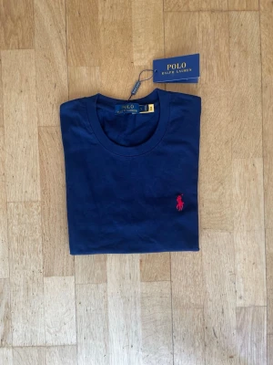 Marinblå t-shirt från Polo Ralph Lauren - Snygg marinblå t-shirt från Polo Ralph Lauren med klassisk röd logga broderad på bröstet. Tillverkad i mjuk bomull och har en normal passform med korta ärmar och rund halsringning. Perfekt för en clean och stilren look. Säg till om ni vill ha fler bilder eller har frågor!👍 kvitto finns inte kvar tyvärr