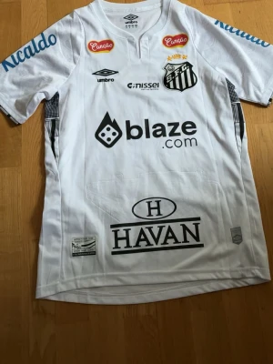 Vit Santos FC Neymar Jr matchtröja - Säljer en vit Santos FC matchtröja från Umbro med Neymar Jr och nummer 10 på ryggen. Tröjan har flera sponsorloggor, klubbmärke på bröstet och svarta detaljer. Perfekt för dig som älskar brasiliansk fotboll och vill ha en ikonisk supportertröja.