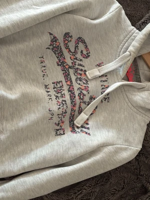 Grå hoodie från Superdry med blommigt tryck - Snygg ljusgrå hoodie från Superdry med blommigt tryck på bröstet och texten 'Vintage Superdry'. Hoodien har dragsko i huvan och ribbade muddar. Perfekt för en avslappnad och trendig stil. Materialet känns mjukt och bekvämt.