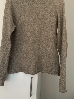 Beige stickad tröja i ullmix - Mysig beige stickad tröja med rund hals och långa ärmar. Tröjan har ribbstickade muddar vid ärmslut och nederkant, vilket ger en klassisk och enkel look. Perfekt för lager på lager under kyliga dagar.