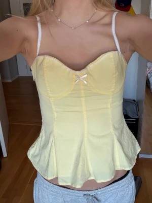 Gul korsettopp med rosett - Söt ljusgul korsettopp med tunna axelband och liten rosett framtill. Toppen har en figurnära passform och lätt utsvängd nederkant som ger en feminin siluett. Perfekt till jeans eller kjol för en trendig look. Använd typ 2 tillfällen men annars som ny. Assnygg på sommarn när man är brun.