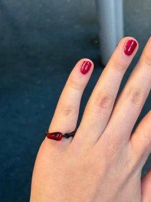 Svart ring med röd pärla - Ring bestående av svart metalltråd. Fäst till ringen är en glansig röd pärla formad som en droppe. Passar perfekt för dig som gillar accessoarer med en unik twist.