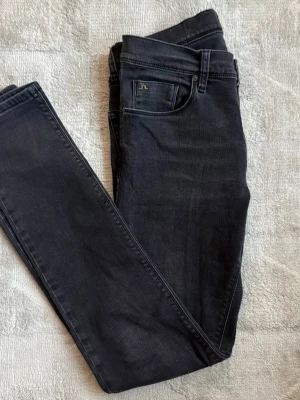 svart slim fit jeans från J.Lindeberg - Snygga svarta slim jeans från J.Lindeberg med klassisk femficksdesign och diskret logga på myntfickan. Jeansen har smal passform och är tillverkade i stretchigt denimtyg för extra komfort. Perfekta för en stilren och modern look.