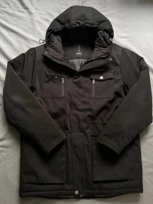 Svart parkas från True North  - Svart parkajacka från True North med huva och flera praktiska fickor framtill. Jackan har dragkedja och tryckknappar, samt justerbar huva. Tillverkad i slitstarkt syntetmaterial och fodrad för extra värme. Perfekt för kalla dagar och nordiskt väder.