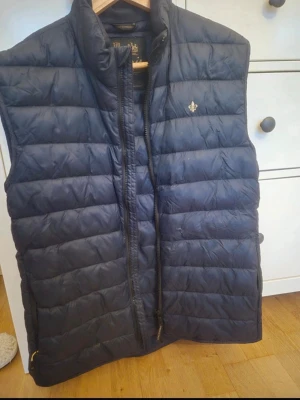 Mörkblå dunväst från Morris - Snygg mörkblå dunväst från Morris med quiltad design och hög krage. Västen har dragkedja framtill och en liten guldfärgad logga på bröstet. Perfekt för lager-på-lager och ger en stilren look till din outfit.