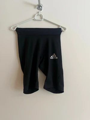 Svarta träningstights från Adidas - Säljer ett par svarta träningstights från Adidas med logga på vänster lår. Tightsen är knälånga och har bred resår i midjan för extra komfort. Tillverkade i stretchigt funktionsmaterial som passar perfekt för gym, löpning eller annan träning.