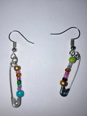Färgglada säkerhetsnål-örhängen med pärlor - Unika örhängen gjorda av silverfärgade säkerhetsnålar dekorerade med små färgglada pärlor i blått, grönt, rosa, guld och orange. Perfekt accessoar för dig som gillar DIY-stil och vill sticka ut med något kreativt och lekfullt.
