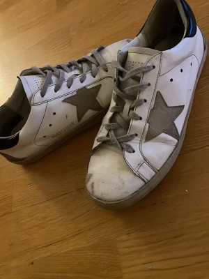 Golden Goose vita sneakers med stjärna - Säljer ett par vita Golden Goose sneakers med grå stjärna på sidan och grå skosnören. Skorna har en låg silhuett och svart detalj på hälen. Tillverkade i läder med slitna detaljer för en cool och avslappnad look.
