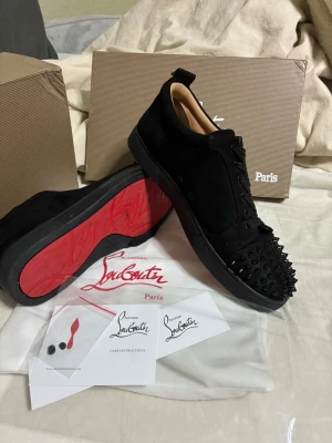Svarta Louboutin sneakers med nitar - Säljer ett par svarta sneakers från Christian Louboutin i mocka med ikoniska röda sulor och coola svarta nitar på tån. Skorna har lågt skaft och klassisk snörning. Perfekta för dig som vill sticka ut med exklusiv design och edgy detaljer.